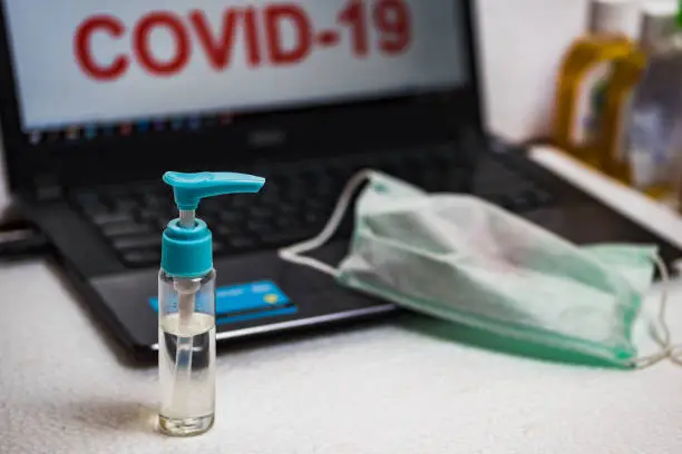 Hand sanitizer bottle and surgical mask on a laptop keyboard that reads COVID-19 on the screen. The concept of preventing office employees from coronavirus disease pandemic.