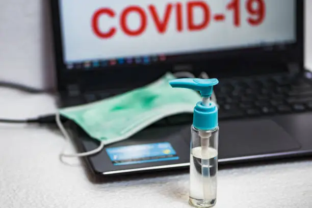 Hand sanitizer bottle and surgical mask on a laptop keyboard that reads COVID-19 on the screen. The concept of preventing office employees from coronavirus disease pandemic.