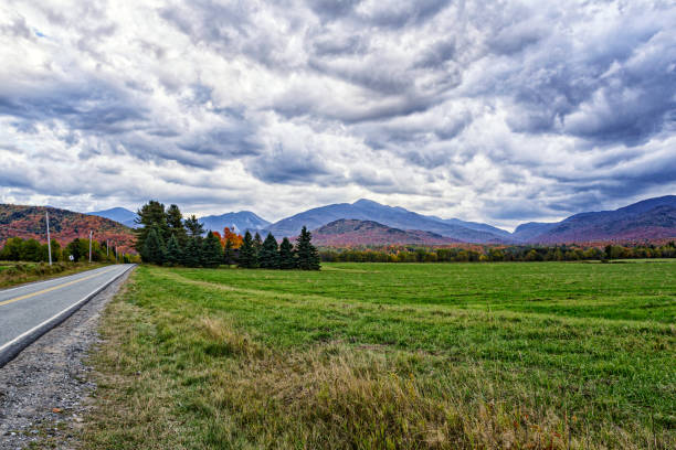 A rural highway in northern New York State runs straight along the edge of an agricultural meadow, with a view of the Adirondack Mountains and a stormy autumn cloudscape in the distance.