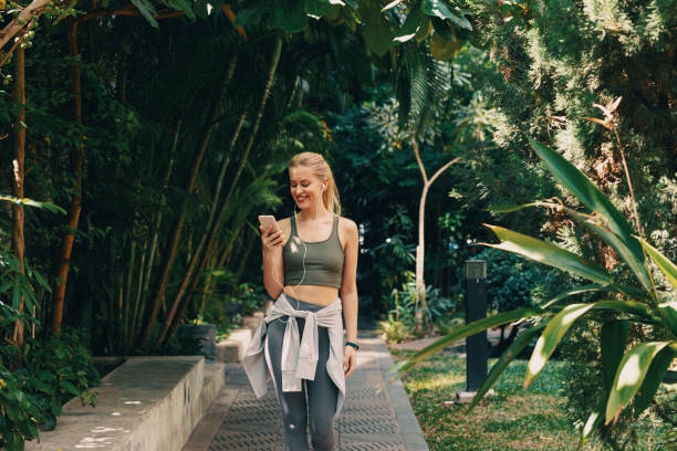entrenamientos al aire libre: mujer rubia feliz en ropa deportiva caminando en el parque mientras escucha música en el teléfono inteligente - tropical climate audio fotografías e imágenes de stock