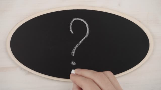 Top view flat lay of a woman's hand drawing a question mark with white chalk on a blackboard