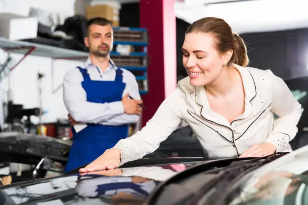 Happy young woman touching surface of repainted car body in auto repair shop