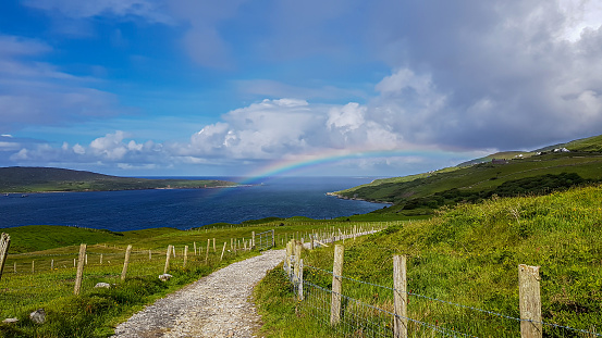 Fences with wooden posts and barbed wire on a dirt trail between the Irish countryside with a rainbow over the Atlantic Ocean, blue sky and white clouds on a spring day in County Galway, Ireland