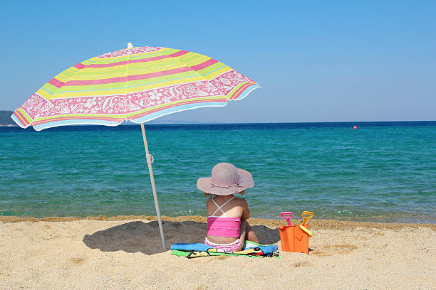 little girl sitting on beach under sunshade stock photo
