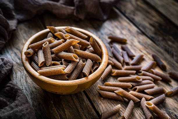 Healthy food: high angle view of a wooden bowl filled with wholegrain pasta shot on rustic wooden table. Some rigatoni are scattered on the table. Predominant color is brown. High resolution 42Mp studio digital capture taken with SONY A7rII and Zeiss Batis 40mm F2.0 CF lens