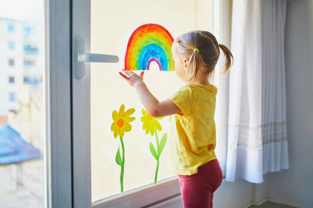 aanbiddelijk peutermeisje dat tekening van regenboog aan vensterglas als teken van hoop vastmaakt - alleen babys stockfoto's en -beelden