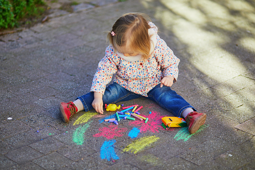 Adorable toddler girl drawing with colorful chalks on asphalt. Outdoor activity and creative games for small kids