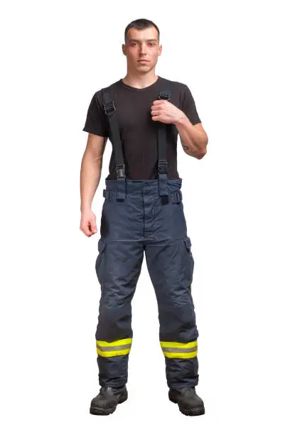 Young firefighter wears black t-shirt standing and pull suspender on his fireproof pants isolated on white background.