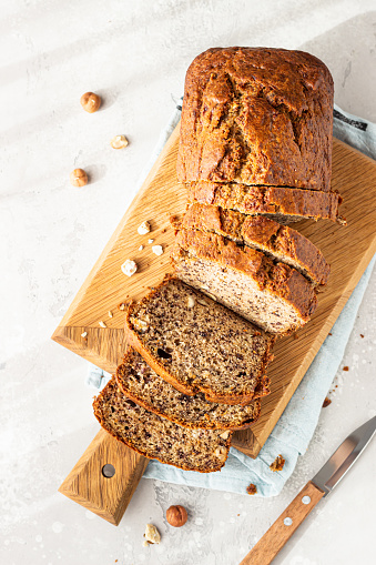 Homemade banana bread or cake with hazelnut on wooden cutting board, light stone background. Trendy soft shadows, atmospheric lighting, selective focus.