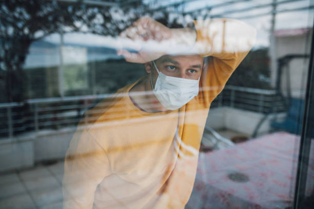 istock Man in quarantine wearing a mask and looking through the window 1216472831