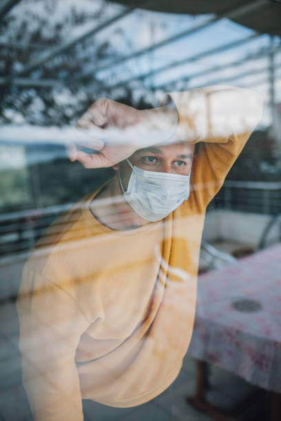 istock Man in quarantine wearing a mask and looking through the window 1216480344