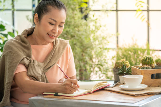 bella donna anziana asiatica che scrive diario o pianifica in quaderno nel giardino della serra di casa. donna in pensione sorridente che si rilassa a casa durante la quarantena. stile di vita e concetto di assistenza sanitaria per gli anziani. - diary writing women human hand foto e immagini stock