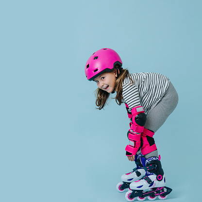 Happy little girl in striped clothes and roller skates with pink protective gear over blue background. She's showing off for a photo, in low groped safe steady position.
