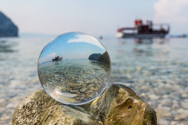 Crystal ball is on stones near the sea. Original upside down view and rounded perspective of the sky, sea and boat. Original and engaging picture.