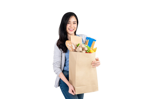 Beautiful smiling Asian woman holding paper shopping bag full of vegetables and groceries isolated on white background