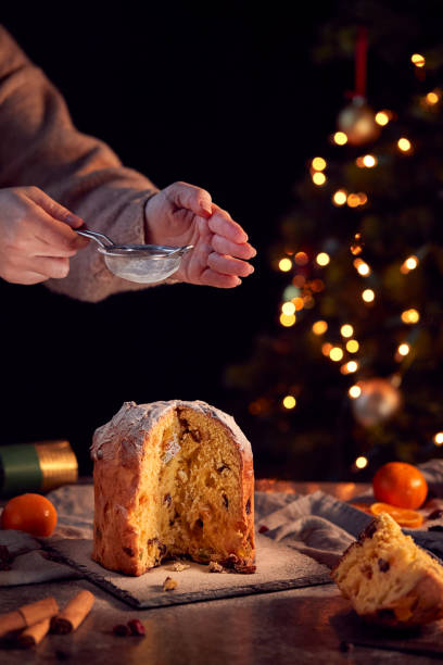 istock Hand Shaking Icing Sugar Over Christmas Panettone On Table Set For Meal With Tree Lights Behind 1216524646