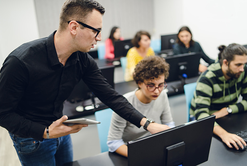 Young people in programming school. Teacher standing in the middle, pointing at computer screen and explaining.