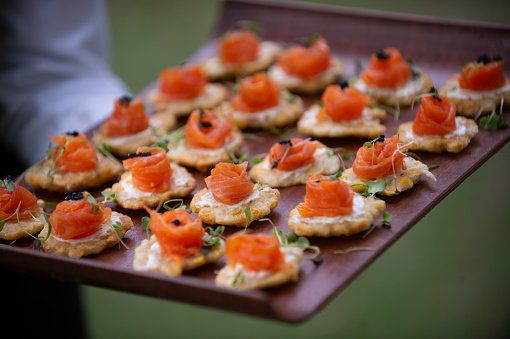 Salmon Appetisers canapes being presented and served at a party with selective focus.  It is being served at a wedding party.  A waiter is holding the wooden tray with canapes. It is salmon with caviar and rocket leaves on cottage cheese on a biscuit that is being served.