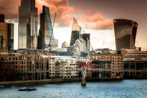 Color image depicting crowds of city workers and tourists walking across Millennium Bridge in central London, UK. Beyond the bridge, the huge modern skyscrapers that make up the contemporary London city skyline dominate the image. It is sunset and the clouds are lent a warm orange glow by the dipping sun.