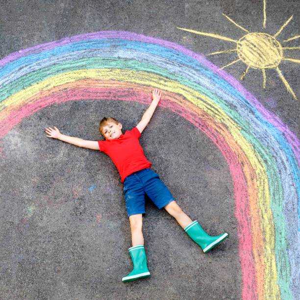 istock Happy kid boy with rainbow painted with colorful chalks on ground during pandemic coronavirus quarantine. Children painting rainbows along with the words Let's all be well. 1216537549