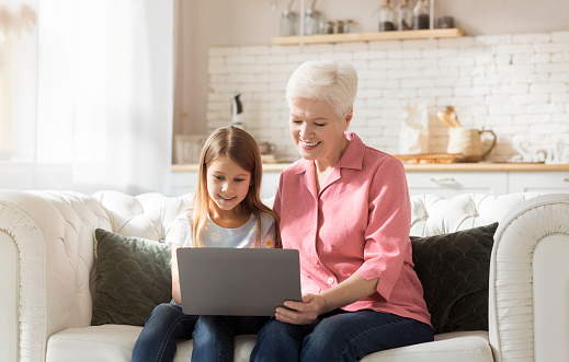 Happy mature woman and her granddaughter using laptop together indoors, playing video games or browsing internet