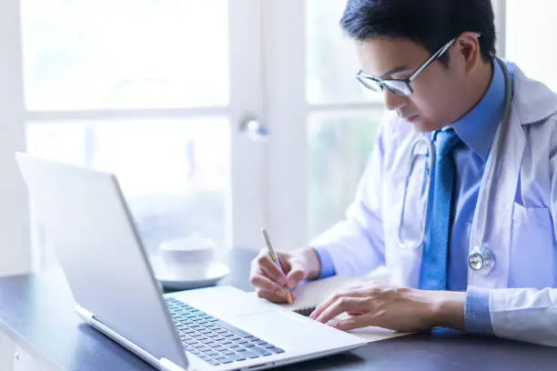 Young asian handsome doctor writing on notebook while using his laptop computer with serious face