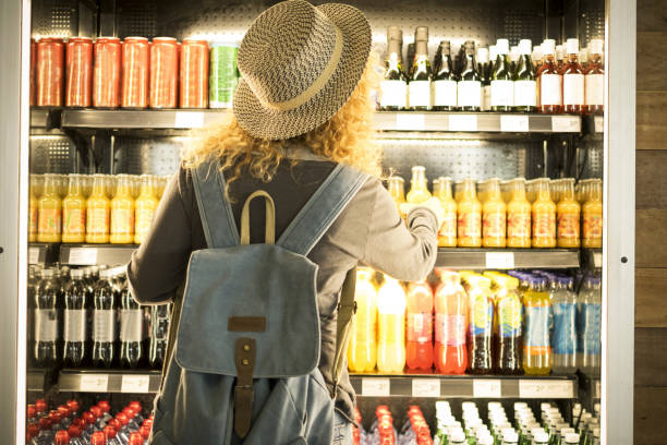 Travel woman viewed from back choosinf beverage in a fresh fridge - airport or station bar concept and traveler passenger choosing drinks - modern lifestyle backpack people buying drinks bottles Travel woman viewed from back choosinf beverage in a fresh fridge - airport or station bar concept and traveler passenger choosing drinks - modern lifestyle backpack people buying drinks bottles non alcoholic beverage photos stock pictures, royalty-free photos & images