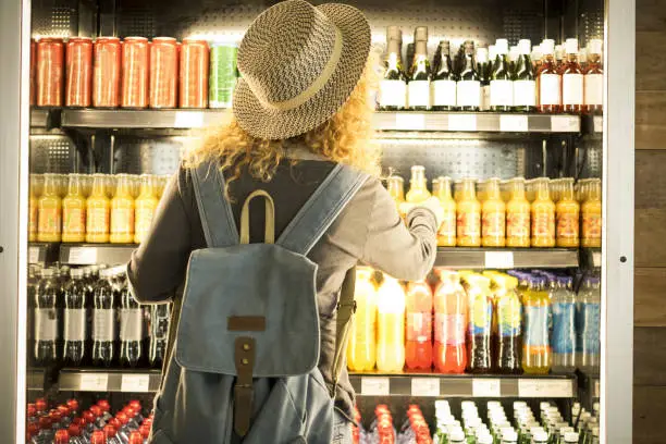Photo of Travel woman viewed from back choosinf beverage in a fresh fridge - airport or station bar concept and traveler passenger choosing drinks - modern lifestyle backpack people buying drinks bottles