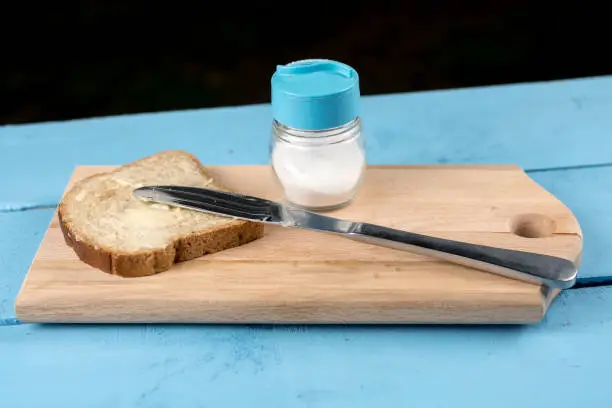Flat lay butter spread on bread with knife and saltshaker served on a wooden board.