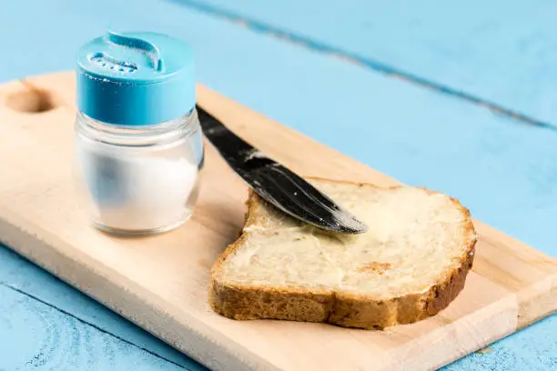 Flat lay butter spread on bread with knife and saltshaker served on a wooden board.