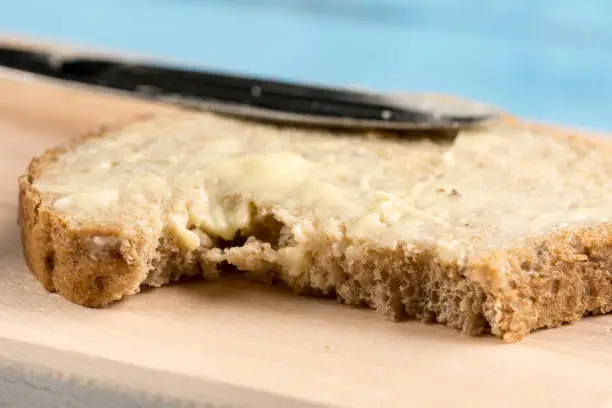 Flat lay butter spread on bread with knife and saltshaker served on a wooden board.