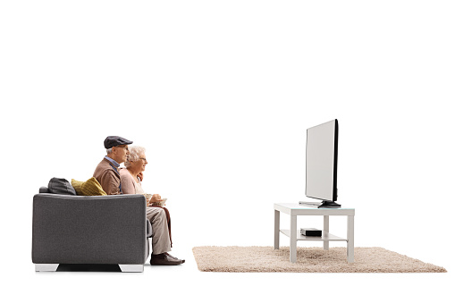 Profile shot of a senior man and woman sitting on a sofa and watching tv isolated on white background