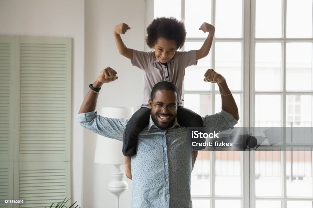 African ethnicity little son sitting on fathers shoulders showing biceps Small son sit on strong dad shoulders showing biceps. African family enjoy activity games at home, healthy fit lifestyle, two superheroes, vitamins for adults and children ad, happy Father Day concept Healthy Lifestyle Stock Photo