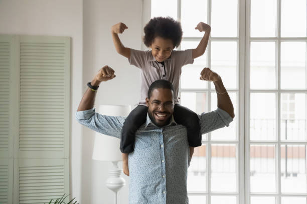 African ethnicity little son sitting on fathers shoulders showing biceps Small son sit on strong dad shoulders showing biceps. African family enjoy activity games at home, healthy fit lifestyle, two superheroes, vitamins for adults and children ad, happy Father Day concept muscular build stock pictures, royalty-free photos & images