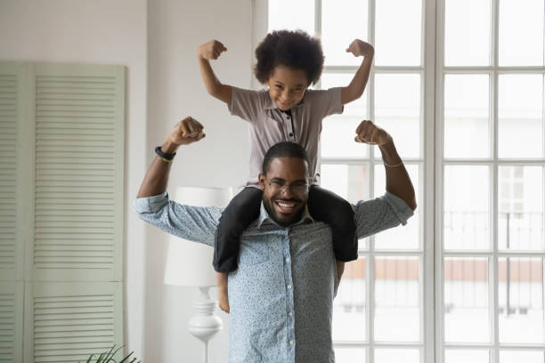 African ethnicity little son sitting on fathers shoulders showing biceps Small son sit on strong dad shoulders showing biceps. African family enjoy activity games at home, healthy fit lifestyle, two superheroes, vitamins for adults and children ad, happy Father Day concept unyielding stock pictures, royalty-free photos & images