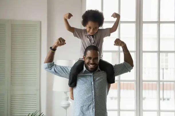 Photo of African ethnicity little son sitting on fathers shoulders showing biceps