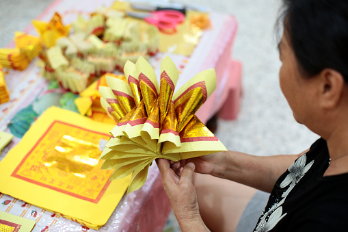 A senior woman is folding gold ingot 'paper money'. It is with a purpose to pray the ancestors with offerings during annual tomb sweeping day at home due lockdown in Malaysia.