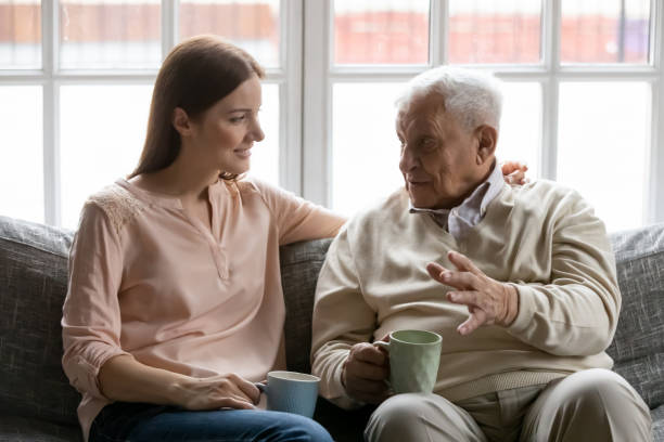 Adult daughter listens to aged father during teatime at home Talk during teatime with aged man, caregiver listens life memories stories of old 75s patient seated on sofa in cozy living room. Provide support care and attention of older generation people concept father daughter 80s stock pictures, royalty-free photos & images