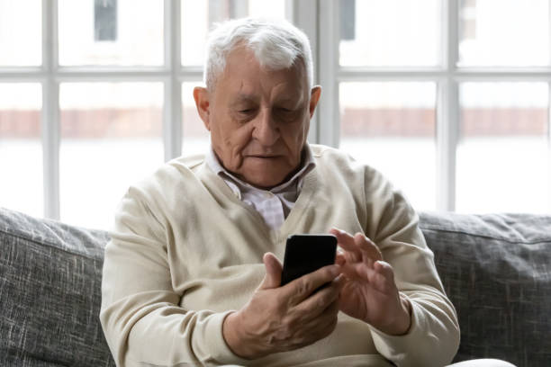 istock Grey-haired 80s old man sitting on couch with smartphone 1216570699