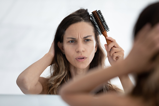 Woman looks in mirror feels dissatisfied by hairstyle, hair condition, holds hairbrush combing hair, hairloss problems vitamin deficiency sign, blow-dryer straightener overuse, need treatment concept
