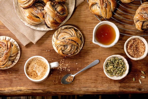 Swedish cardamom buns Kanelbulle Traditional Swedish cardamom sweet buns Kanelbulle on cooling rack, ingredients in ceramic bowl above, cup of coffee on wooden table. Flat lay, space kanelbulle stock pictures, royalty-free photos & images
