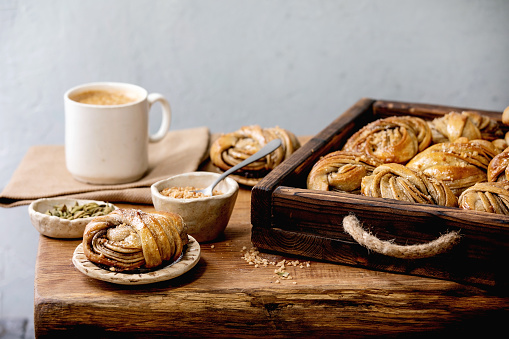 Traditional Swedish cardamom sweet buns Kanelbulle in wooden tray, cup of coffee, ingredients in ceramic bowl above on wooden table.