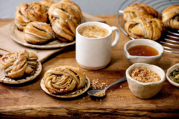 Swedish cardamom buns Kanelbulle Traditional Swedish cardamom sweet buns Kanelbulle on cooling rack, ingredients in ceramic bowl above, cup of coffee on wooden table. kanelbulle stock pictures, royalty-free photos & images