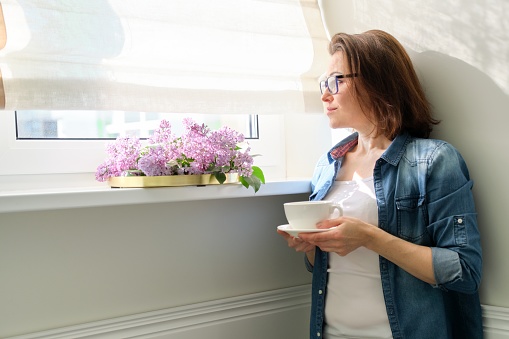 Beautiful mature woman at home with flowers, smiling female standing near window with cup of tea and bouquet of lilacs, copy space