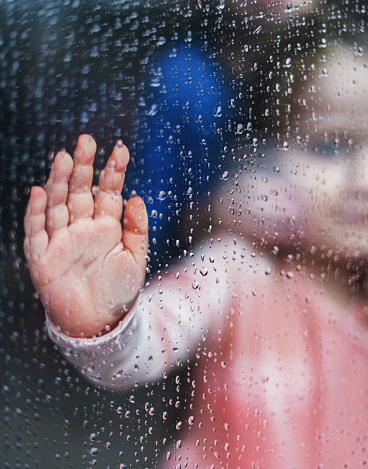 A three year old girl places her hand on a rain soaked window.