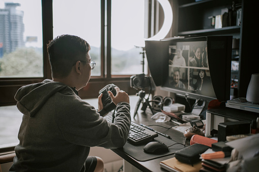 an asian chinese male at his home office vlogging / online blogging e - learning with computer desktop PC, keyboard, computer mouse, monitor scree, Bluetooth speaker, DSLR camera with tripod beside window