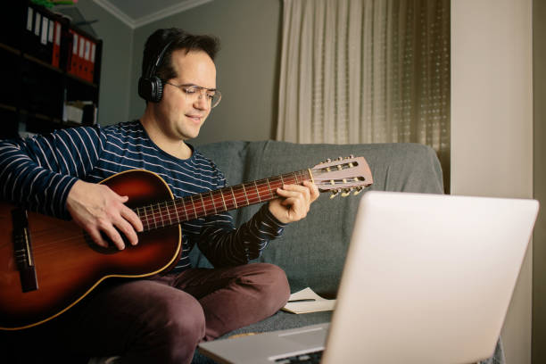 istock Boy doing an online course to learn how to play the classical guitar. He is sitting on the sofa at home, playing the guitar in front of the laptop while listening to the lessons 1216600954