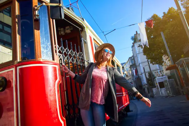 Photo of Beautiful young girl tourist in a hat poses in front of Taksim tram at popular Istiklal street in Beyoglu, Istanbul, Turkey