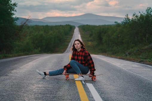 Young Caucasian woman sitting on  longboard   on the road in Finland