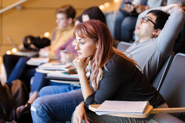College age students listen to lecture; one student sleeps A group of college age students listen to the lecture. One male student sleeps with his arms above his head. male college student sleeping through lecture stock pictures, royalty-free photos & images